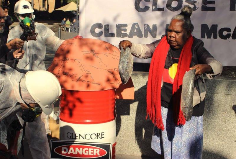 Elder Nancy Yukuwal McDinny with protesters in gas masks and protective gear, slinging raw fish into barrels labelled ‘Glencore’. 