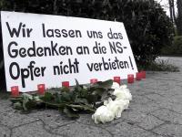 Blumen, Kerzen und Schild mit der Aufschrift "Wir lassen uns das Gedenken an die NS-Opfer nicht verbieten!" am Fuße der Einfahrt zum AWO Psychiatriezentrum