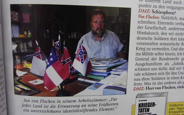 Jan von Flocken posiert für ein Interview der “Deutschen Militärzeitschrift” mit Fahnen rassistischer Regime auf seinem Schreibtisch
