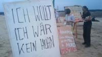 Im Vordergrund: Ein Schild mit der Aufschrift "Ich wollt ich wär kein Huhn", im Hintergrund: Tierschützer protestieren gegen den Bau einer Hähnchenmastanlage.
